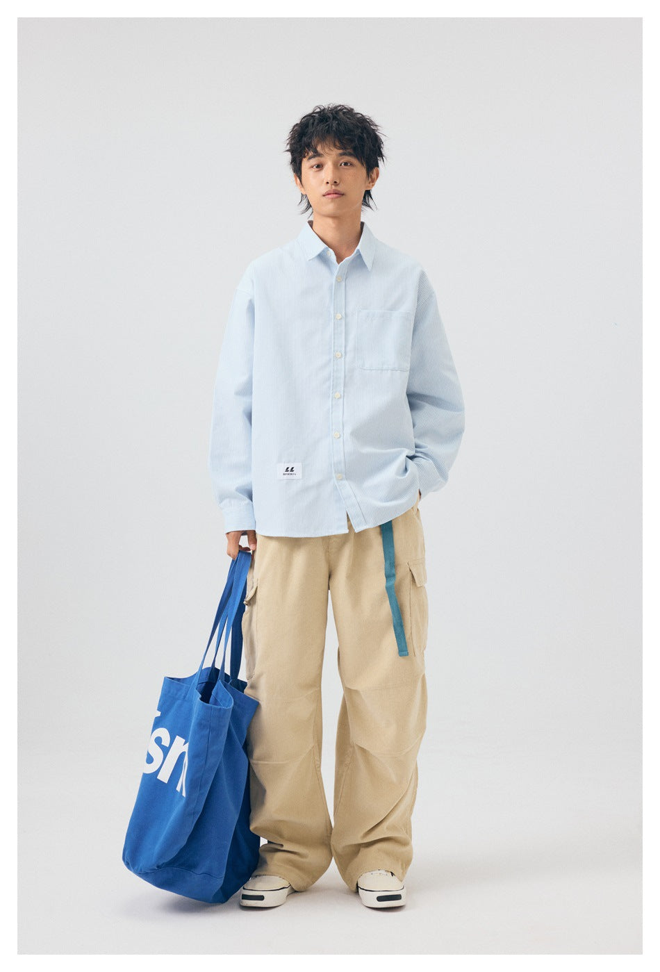 Model wearing light blue striped button-up shirt with khaki cargo pants and holding a blue tote bag