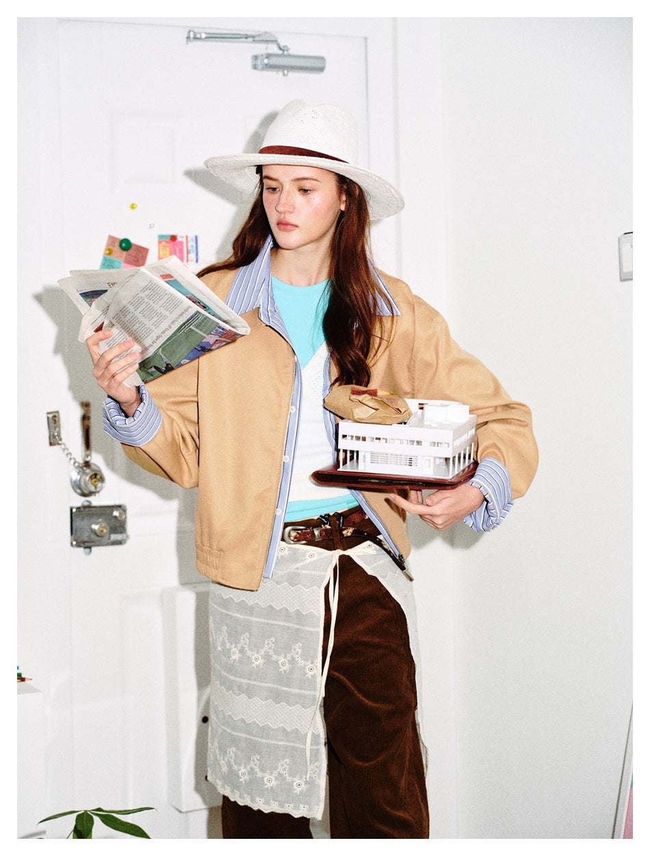 Model wearing an open tan jacket with blue striped collar and cuffs, holding a newspaper and a model building.