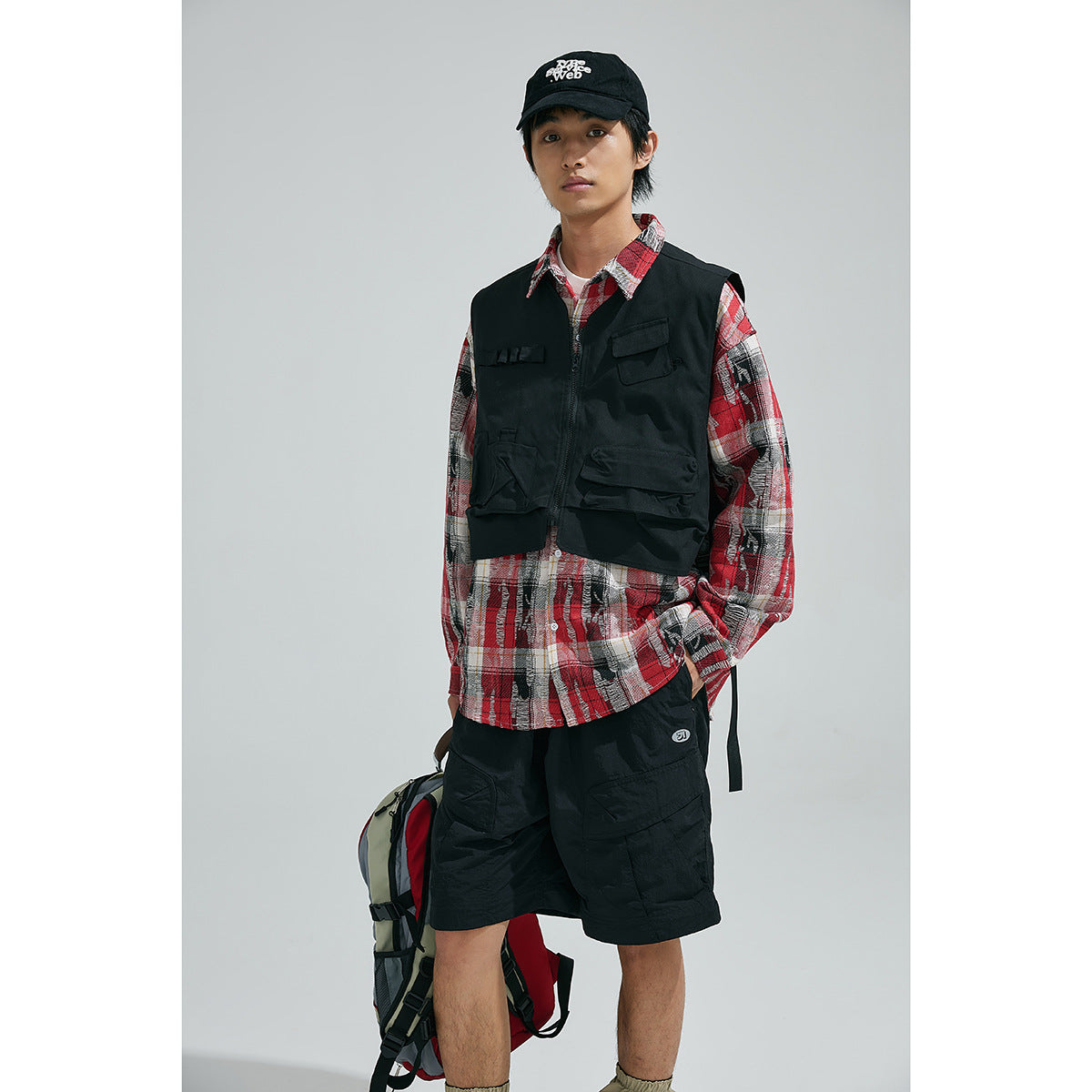 Model wearing a black pocketed utility vest over a red plaid shirt and black shorts.