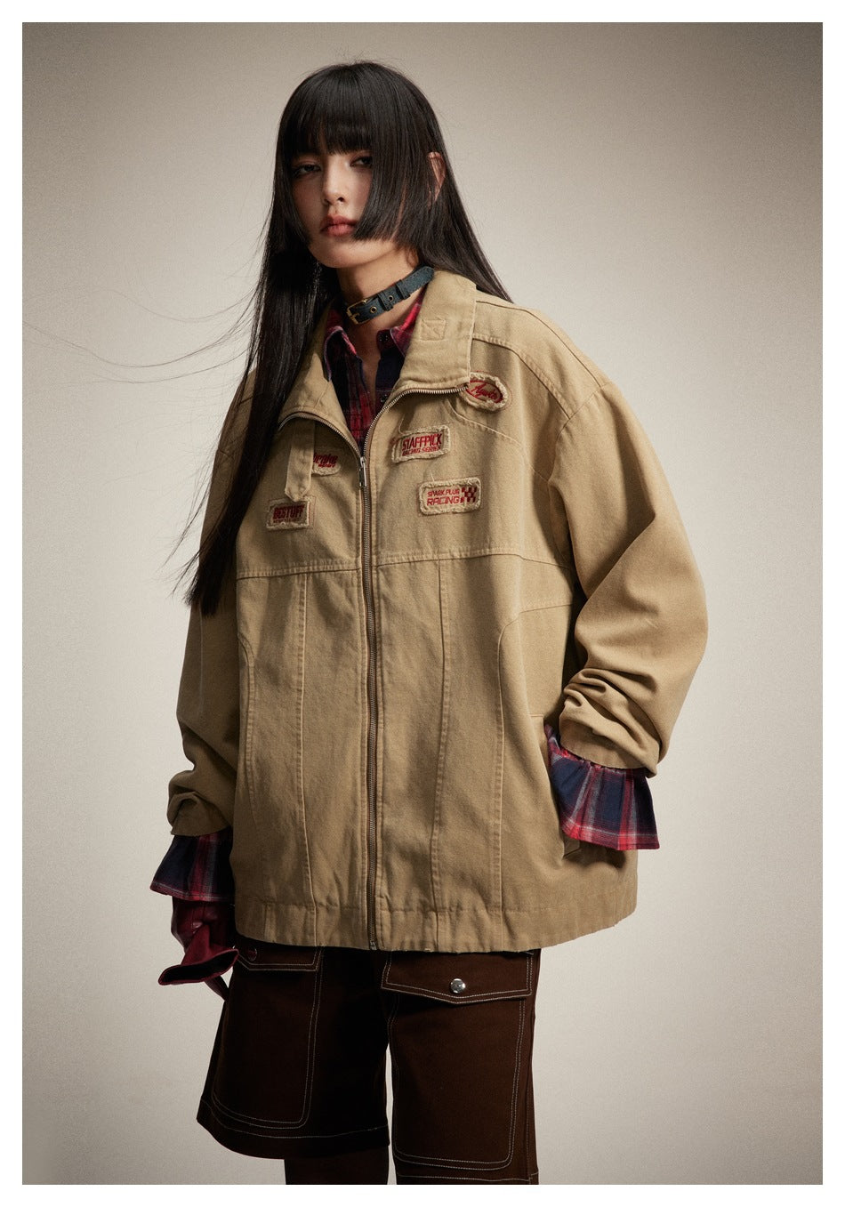 Model wearing a tan zip jacket with embroidered patches, plaid cuffs, a red plaid shirt, and dark shorts.