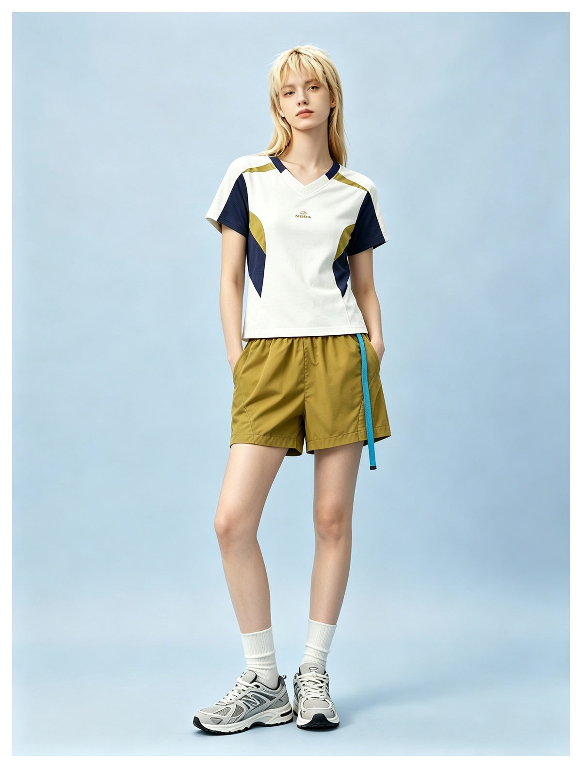 Model wearing yellow shorts with turquoise belt, a color-block tee, white socks and gray sneakers against a blue backdrop