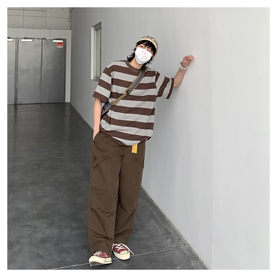 Person posing in a brown and gray striped T-shirt and loose brown cargo pants, wearing a cap and crossbody bag.