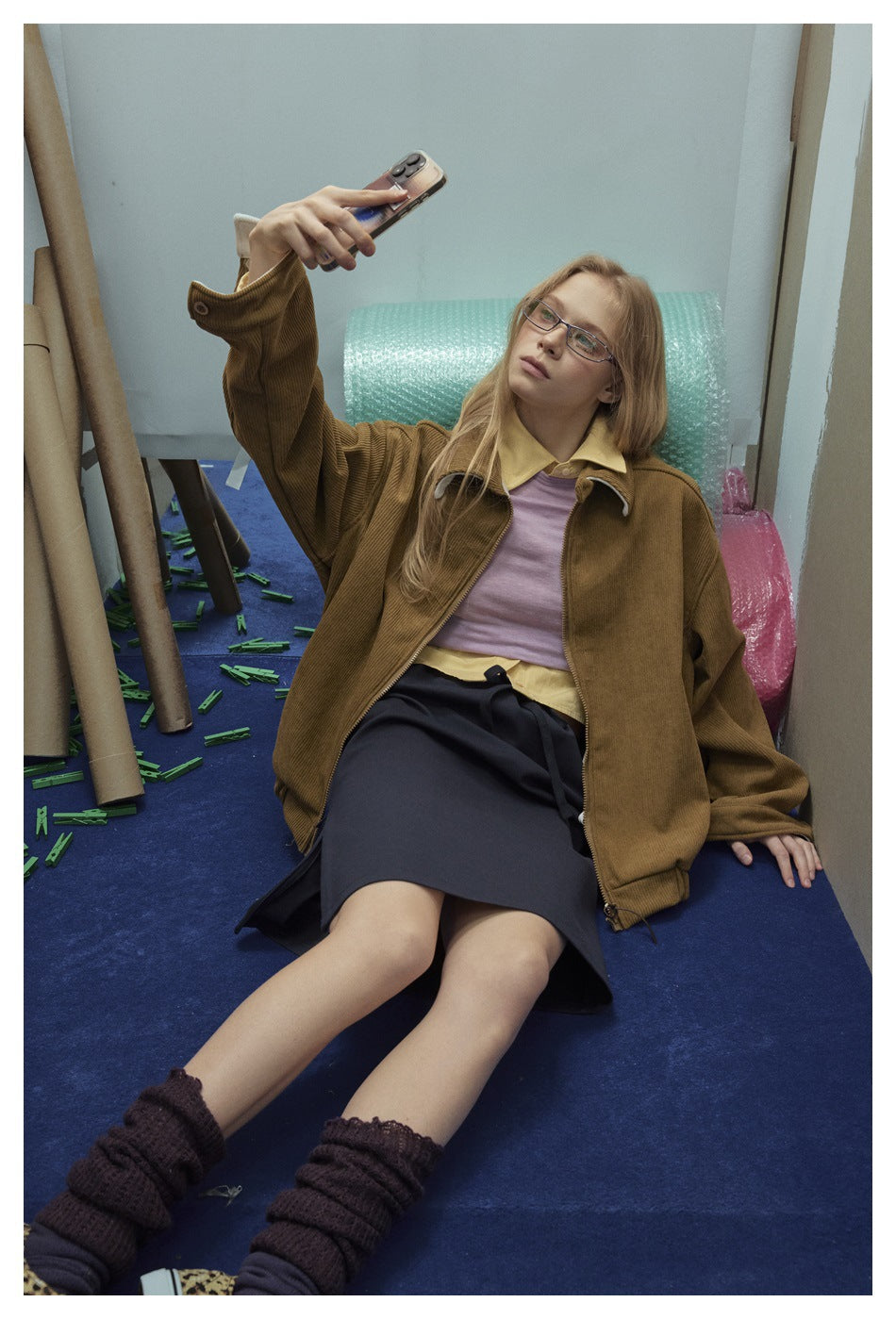 Model seated in a brown ribbed jacket over layered tops and a dark skirt, raising a phone.