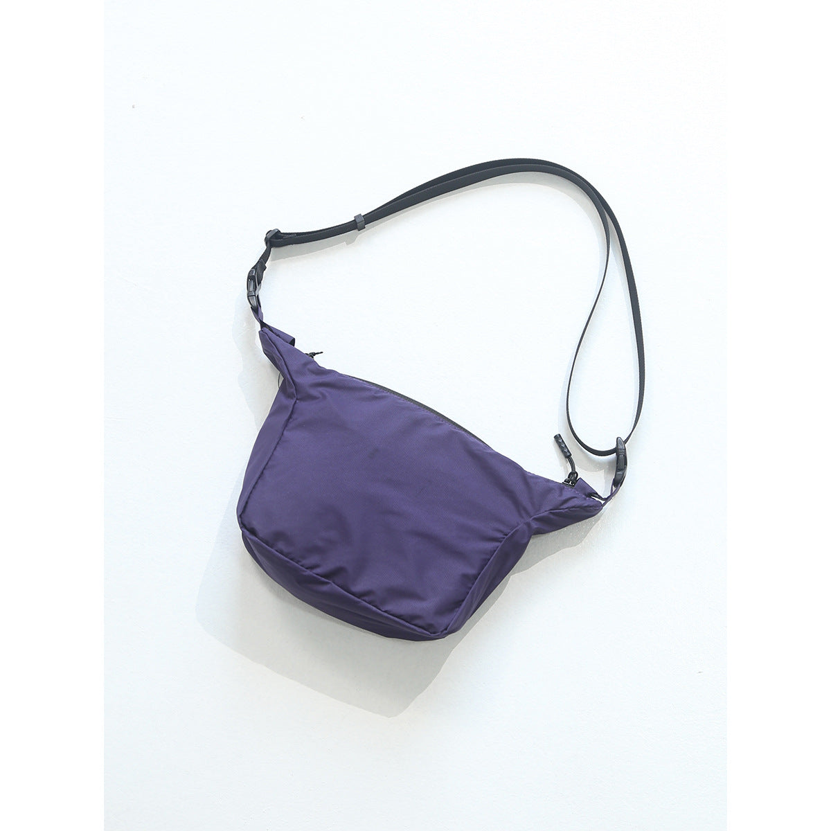 Purple crossbody bag with black adjustable strap, buckle and zipper pull on a light background