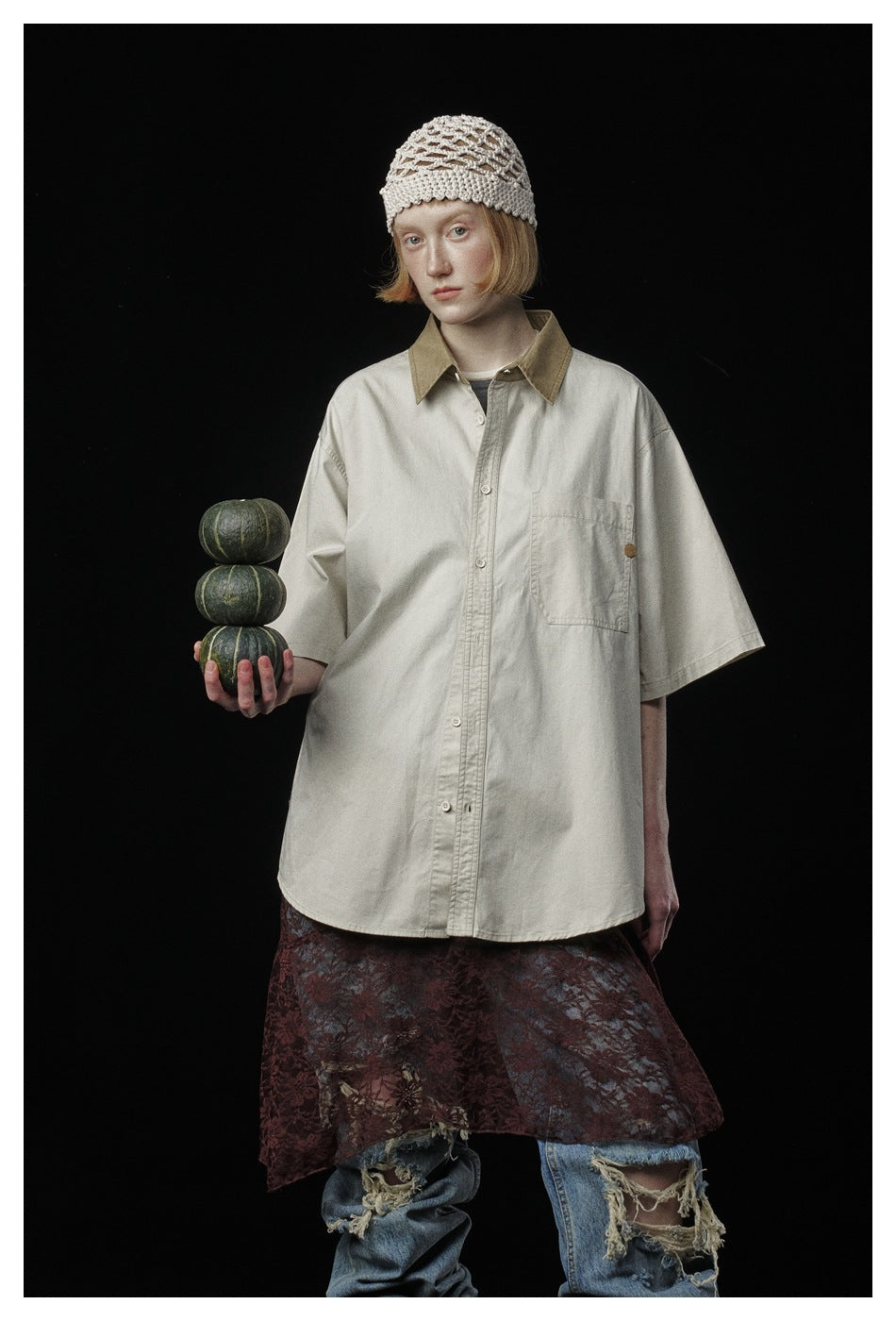 Front view of a model wearing an oversized cream shirt with a tan collar and chest pocket, holding three green gourds.