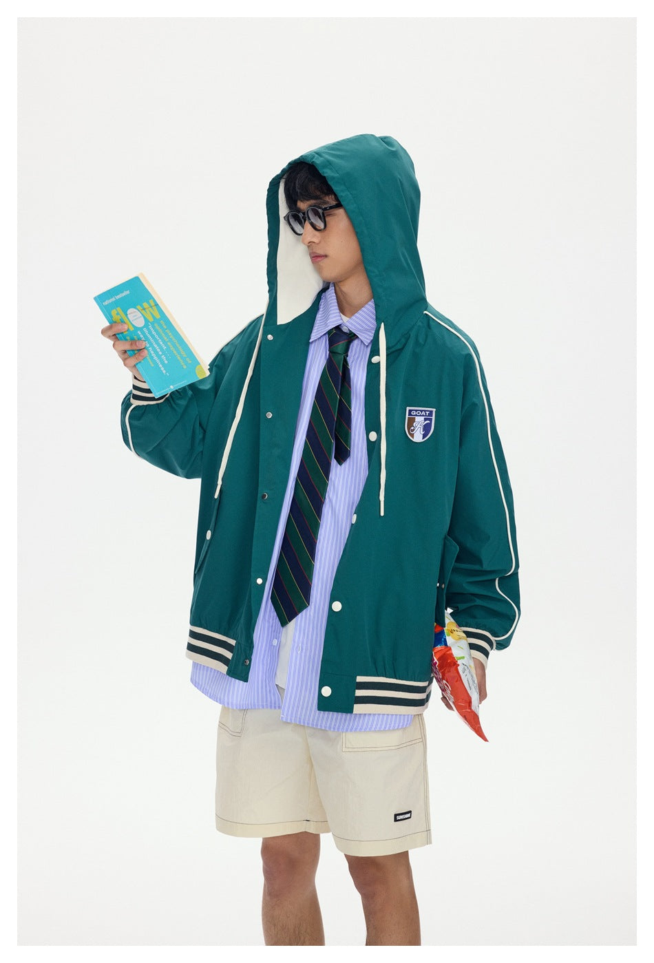 Model in a teal hooded jacket with cream piping and striped trim, holding a book against a white background.