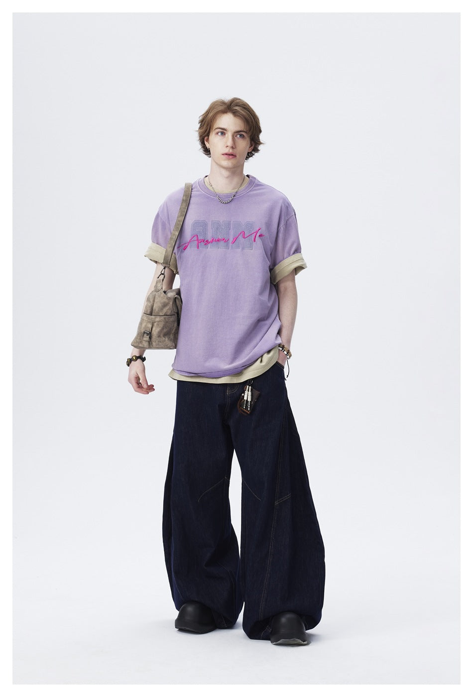 Model wearing a purple ANM Answer Me T-shirt over a khaki tee with wide-leg dark jeans and a shoulder bag