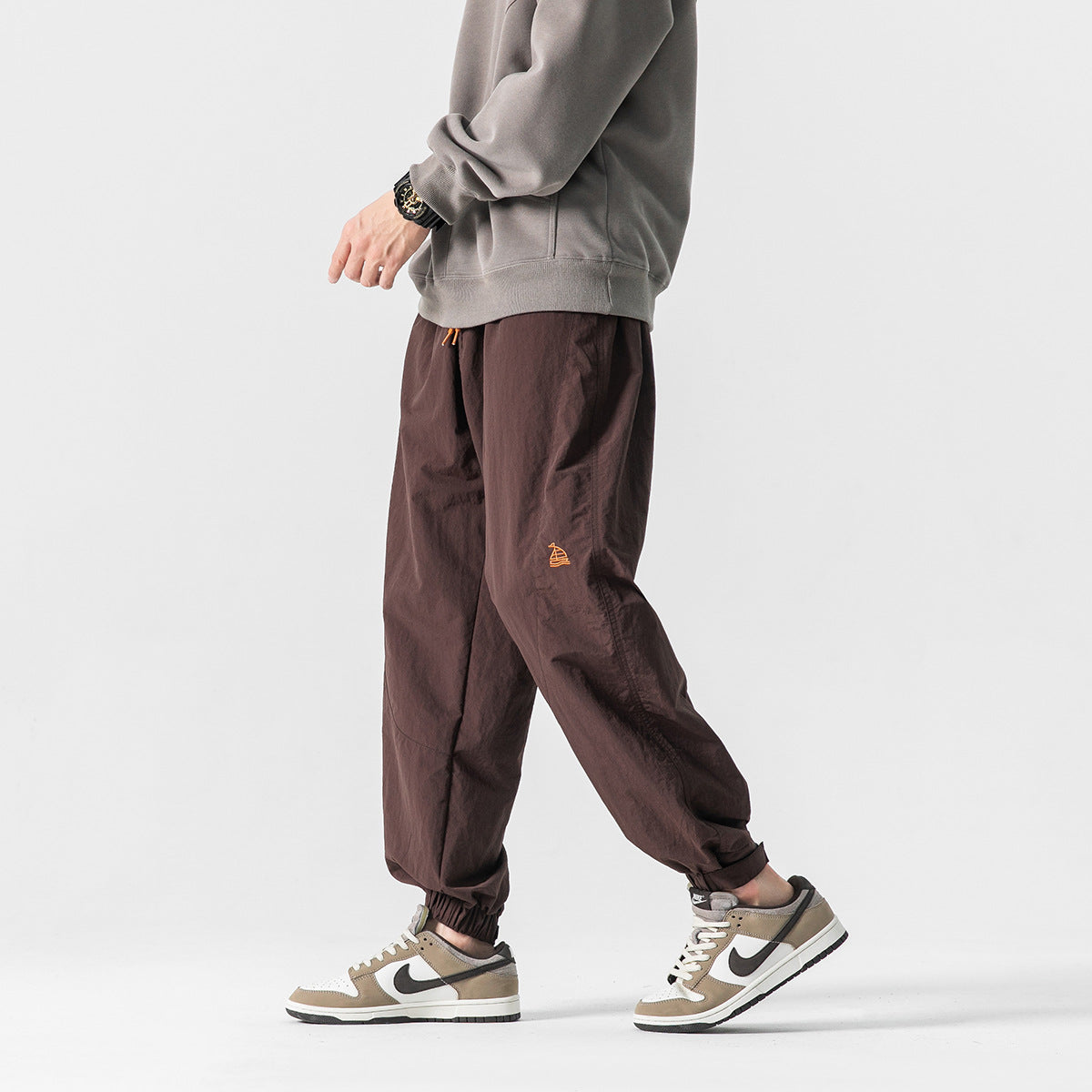 Model wearing dark brown loose jogger pants with elastic cuffs and a small orange emblem, shown at an angle.
