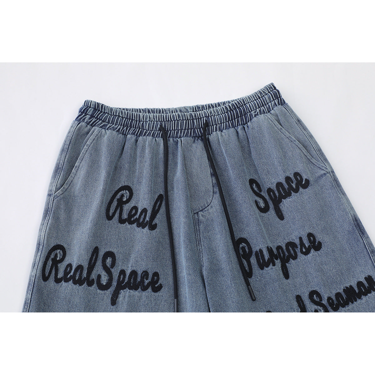 Close-up of blue denim shorts showing an elastic waistband, black drawstrings, pockets, and black script lettering.