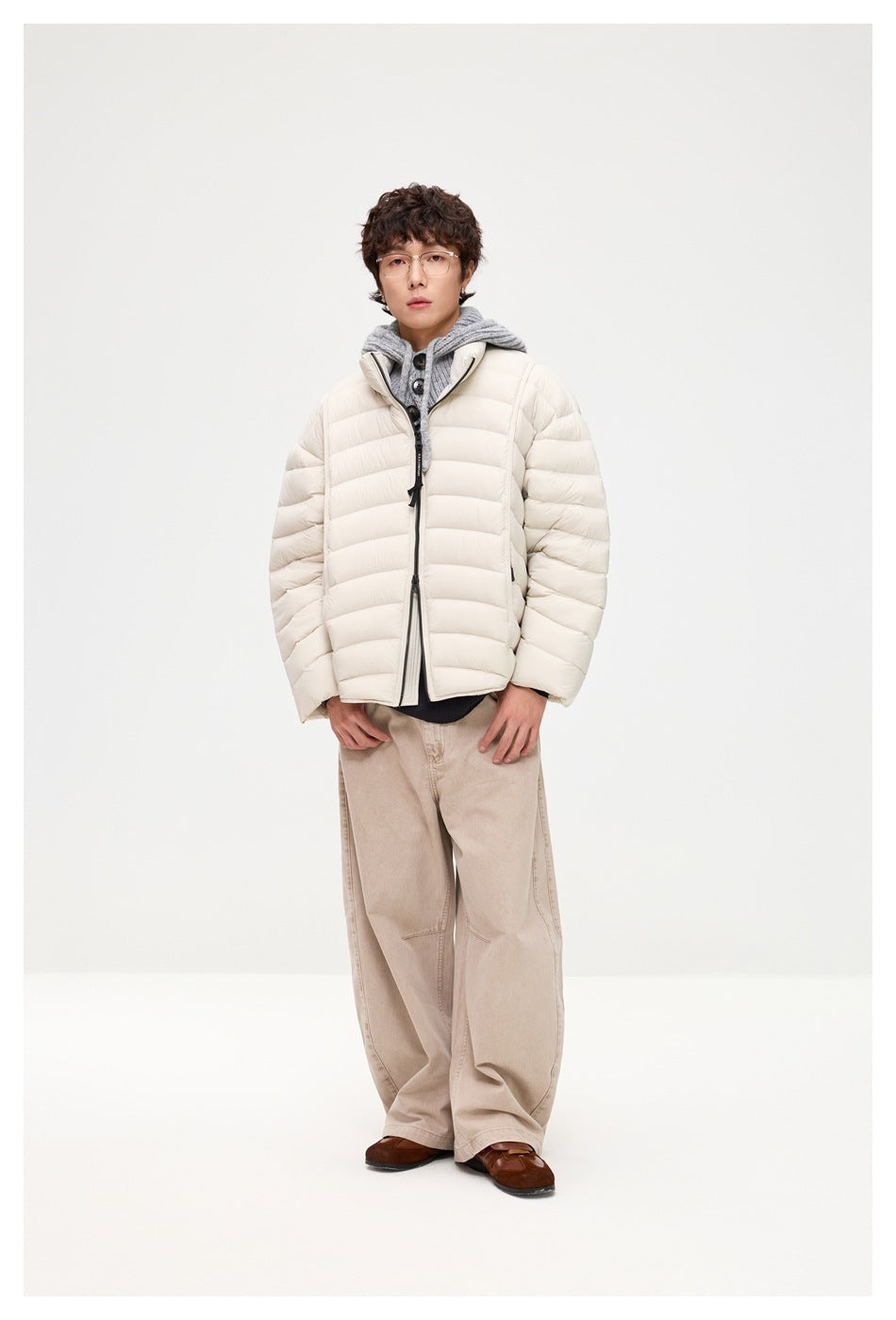 Man wearing a cream quilted puffer jacket with gray hooded knit and wide beige trousers, hands near pockets