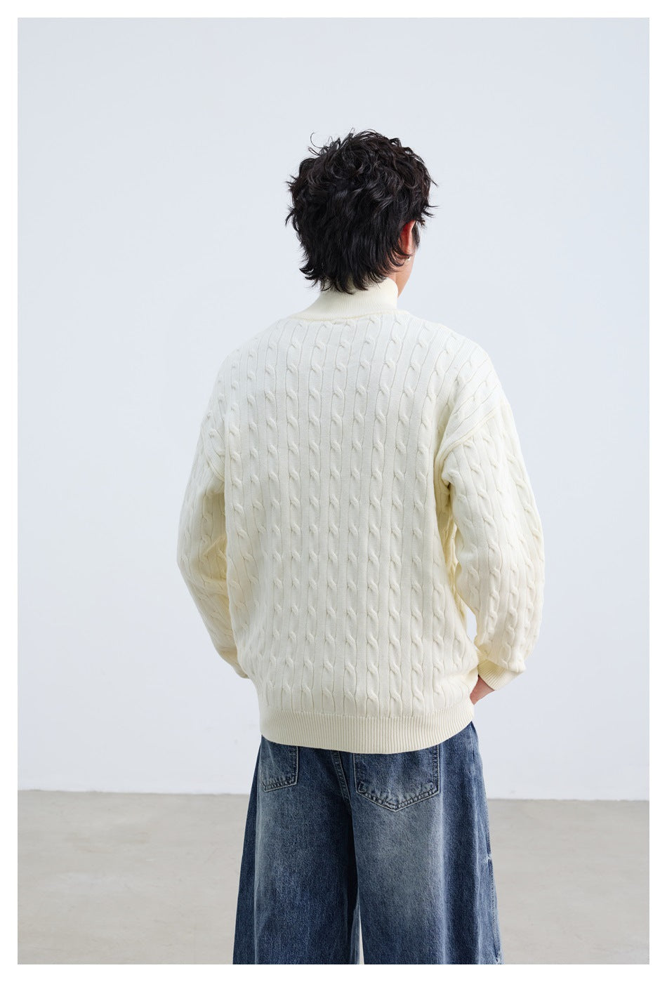 Rear view of a model wearing an ivory cable-knit sweater with ribbed cuffs and hem, paired with blue jeans.