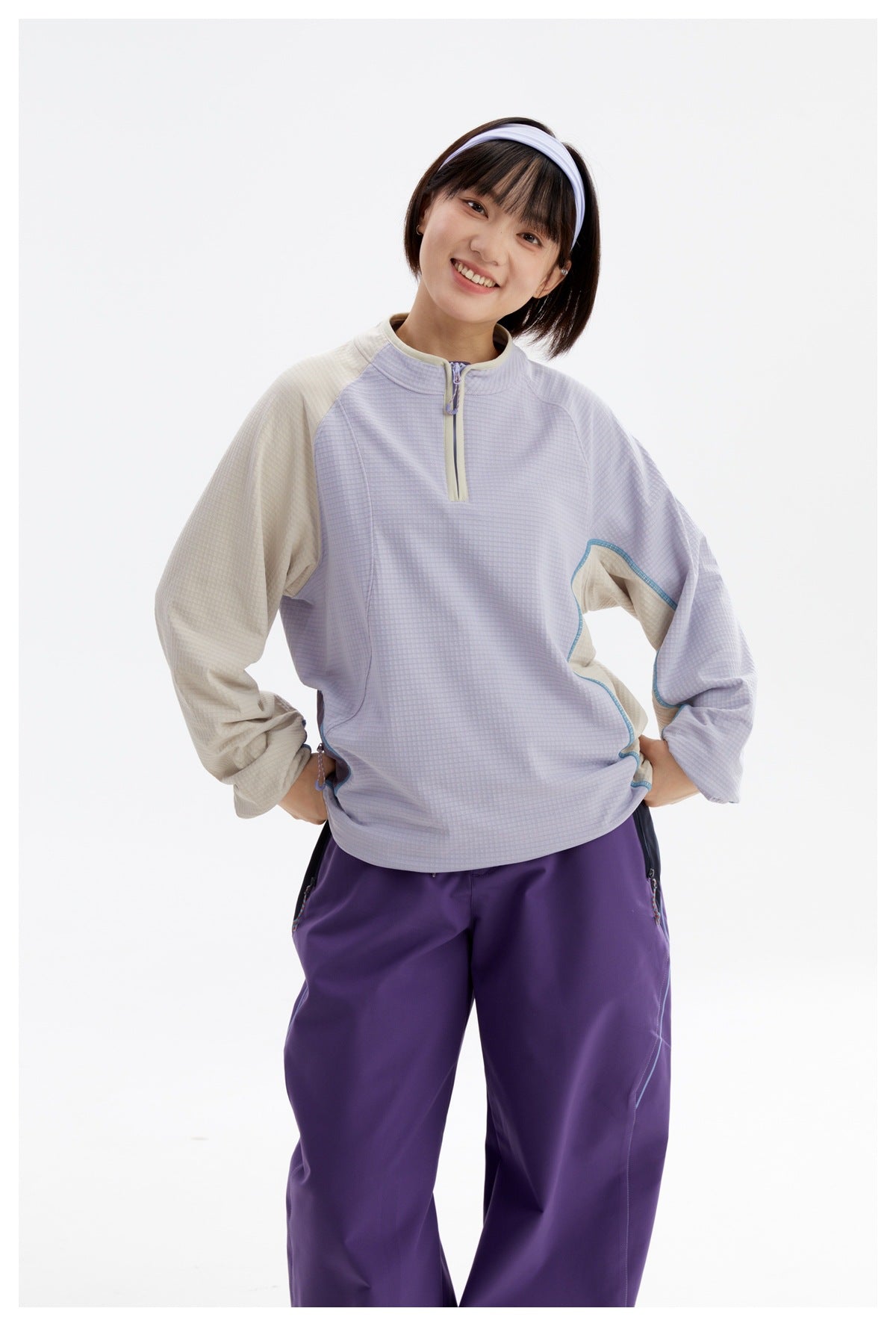 Smiling model wearing a lavender half-zip pullover with cream sleeves and loose purple pants.