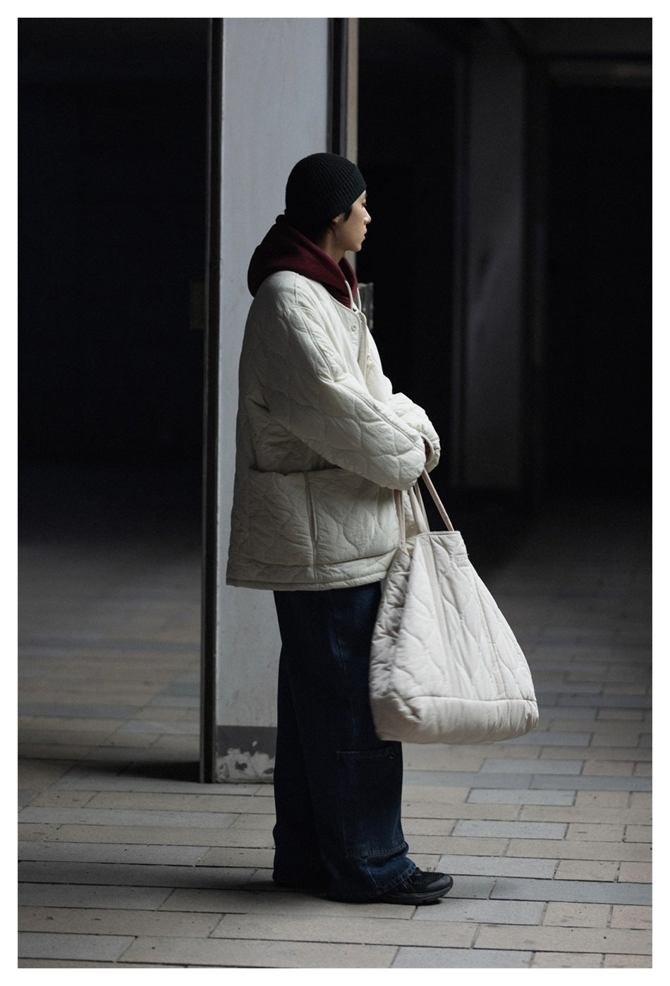 Side view of a model wearing an ivory quilted jacket with dark jeans and a matching tote.