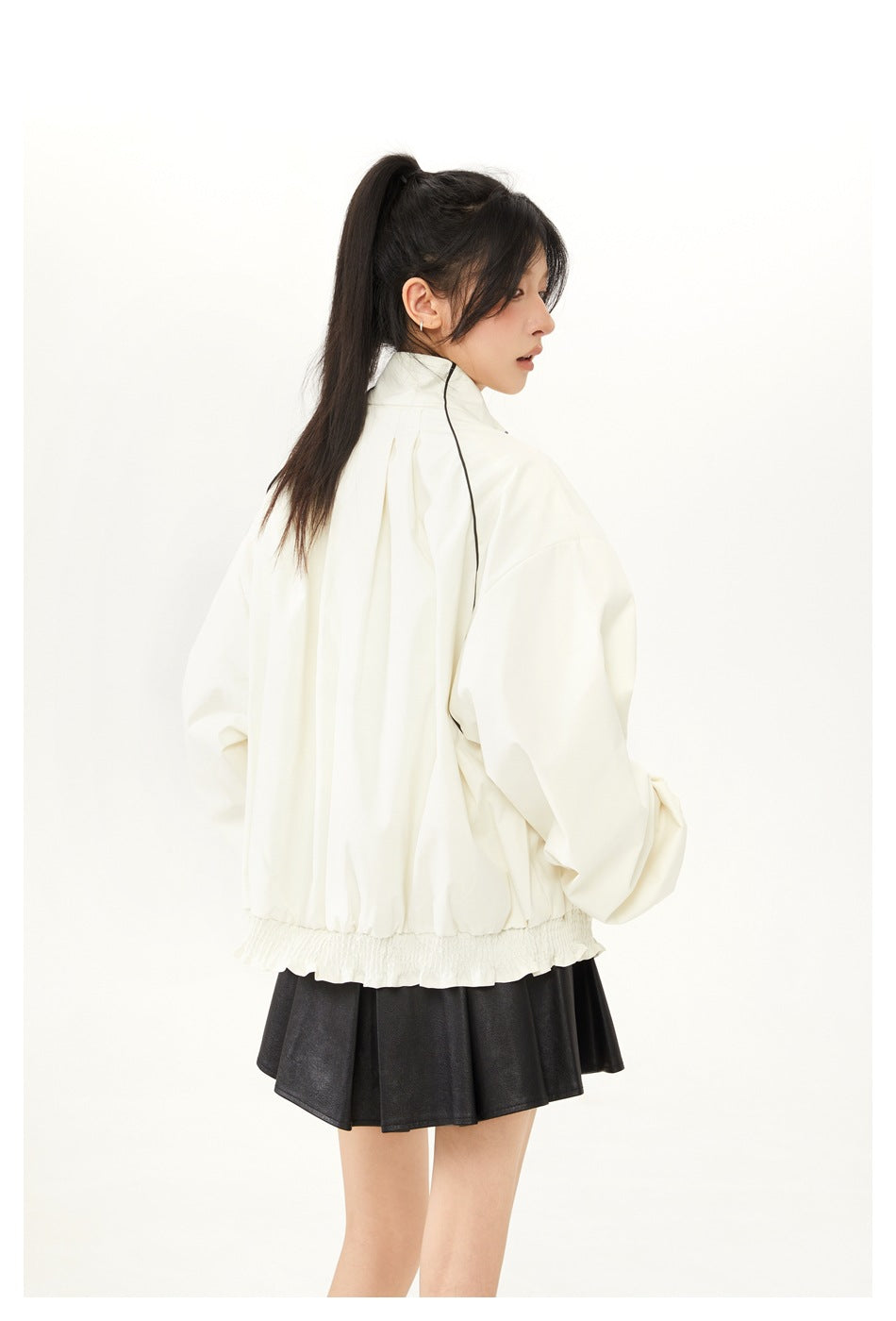 Back view of a model wearing an ivory jacket with black shoulder piping, voluminous sleeves and a gathered hem.