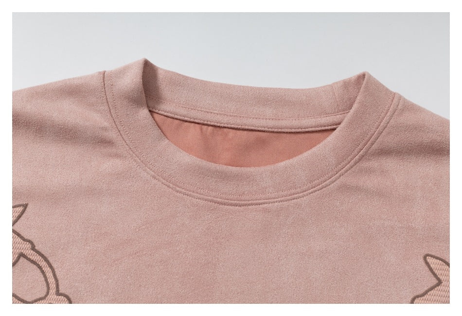 Close-up of the crew neckline and upper chest of a dusty pink T-shirt, with partial outlined embroidery at the edges.