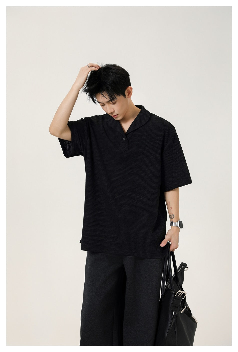 Model wearing an oversized black textured polo with a notched collar, paired with dark trousers and carrying a black bag.