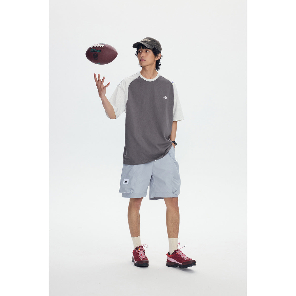 Full-length model wearing a charcoal-and-light-gray raglan T-shirt and pale blue shorts while tossing a football.