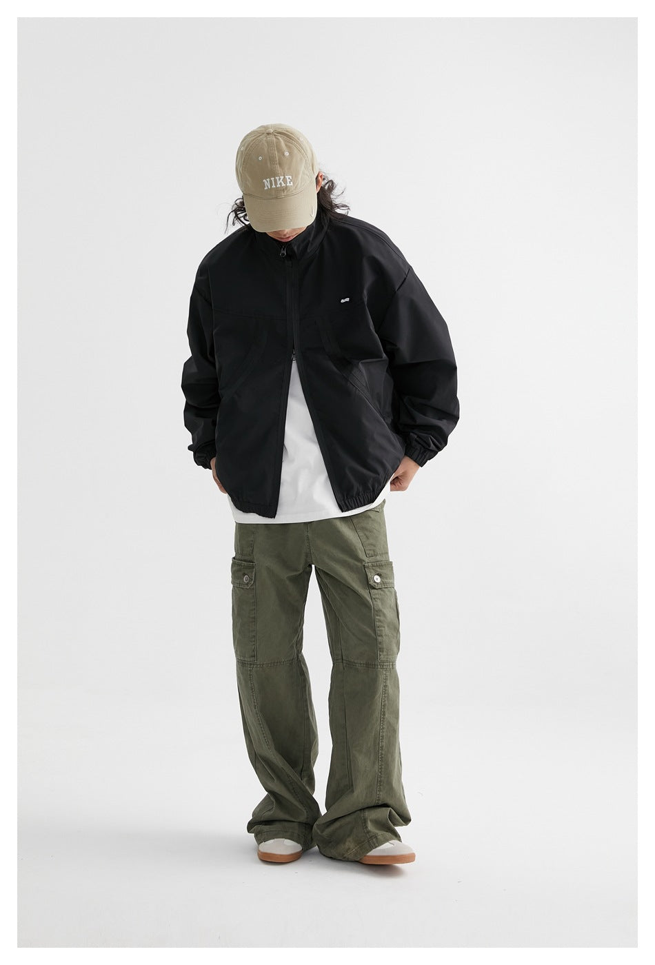 Model with head tilted down wearing the black zip jacket open over a white tee and olive cargo pants