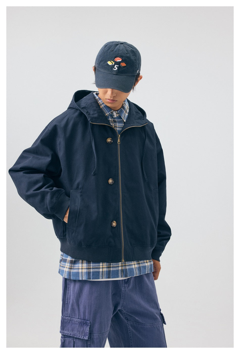 Model wearing the navy hooded jacket zipped closed, showing its front pockets, buttons and central zipper.