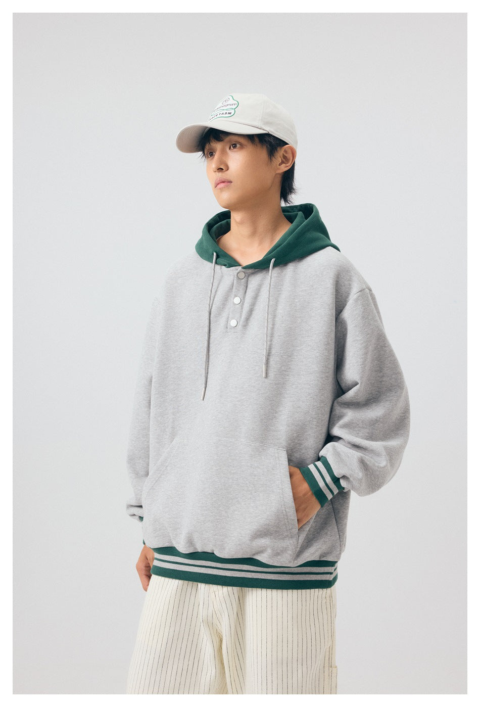Model wearing a light gray pullover hoodie with a dark green hood, snap-button placket, and striped trim.