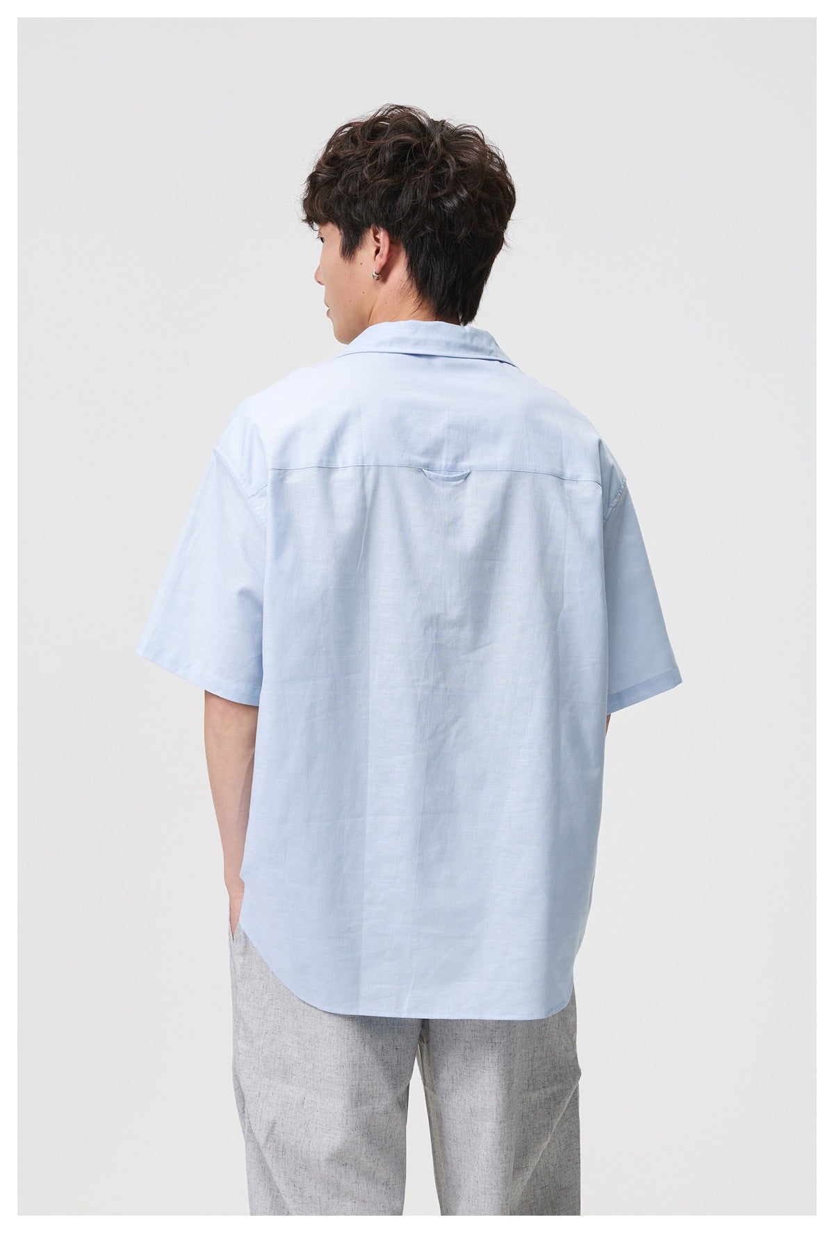 Back view of a model wearing a light blue short-sleeve shirt with a horizontal yoke seam and a small loop detail.