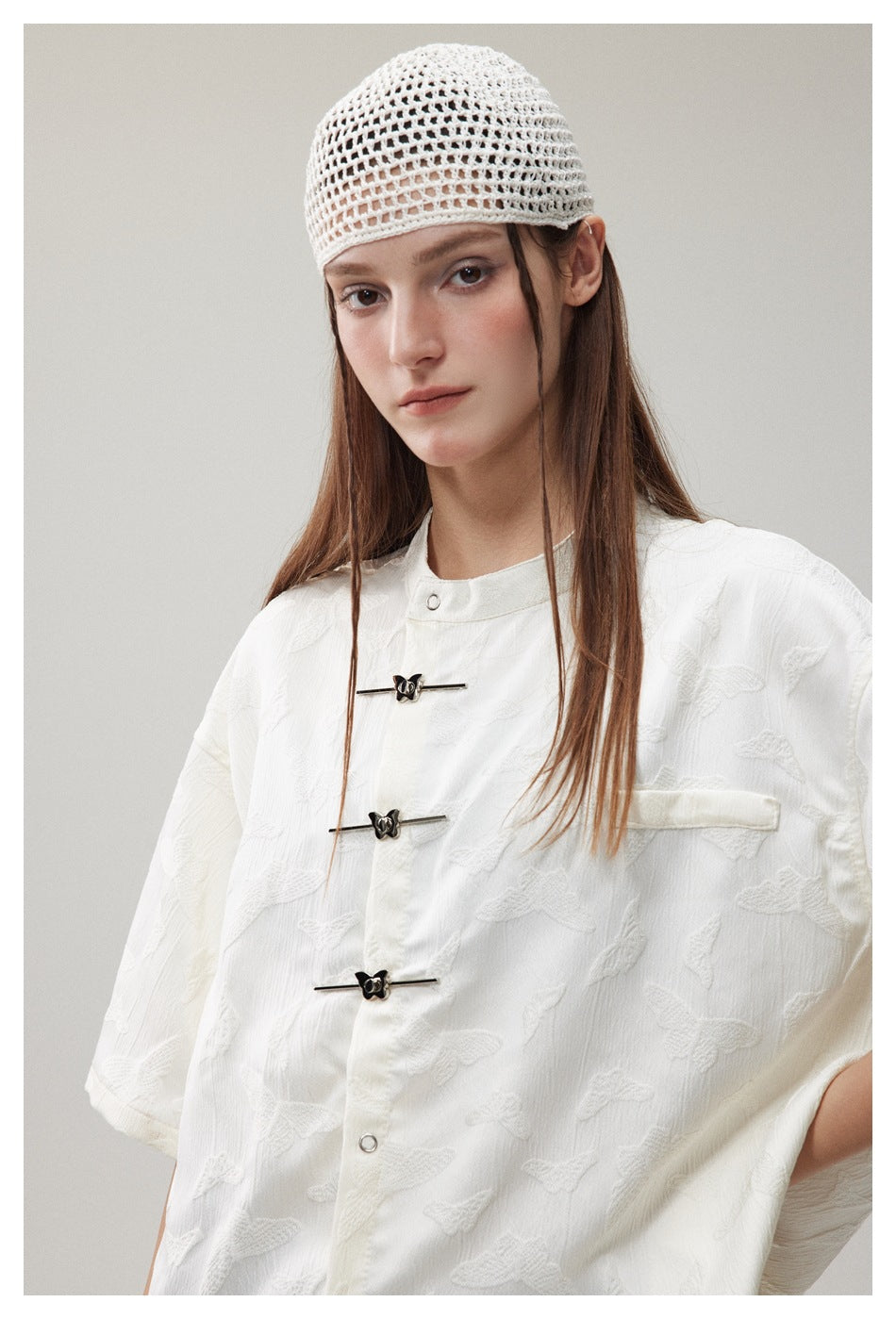 Close-up of a white textured shirt with butterfly-like motifs, three dark frog closures and a chest pocket, worn with a