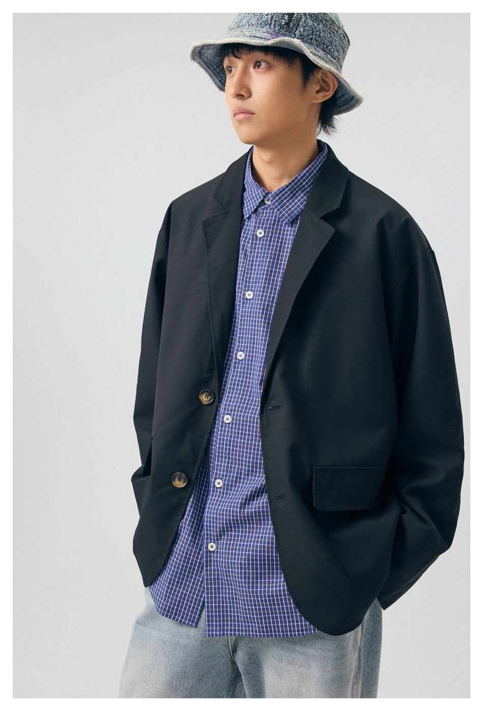 Model wearing a black oversized blazer over a blue checked shirt and light jeans; front view with a denim bucket hat.