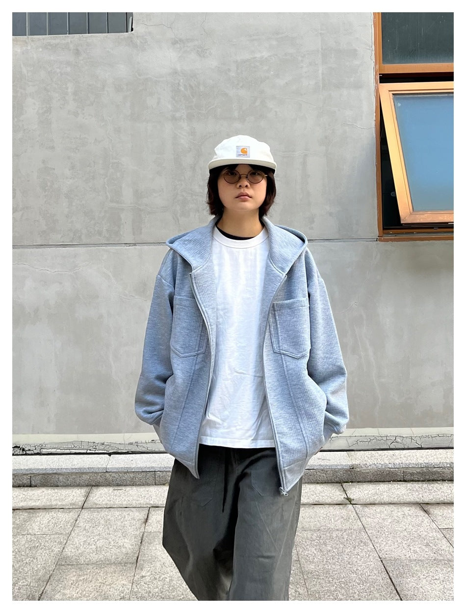 Person wearing an open light gray hooded zip jacket over a white shirt and loose dark pants.