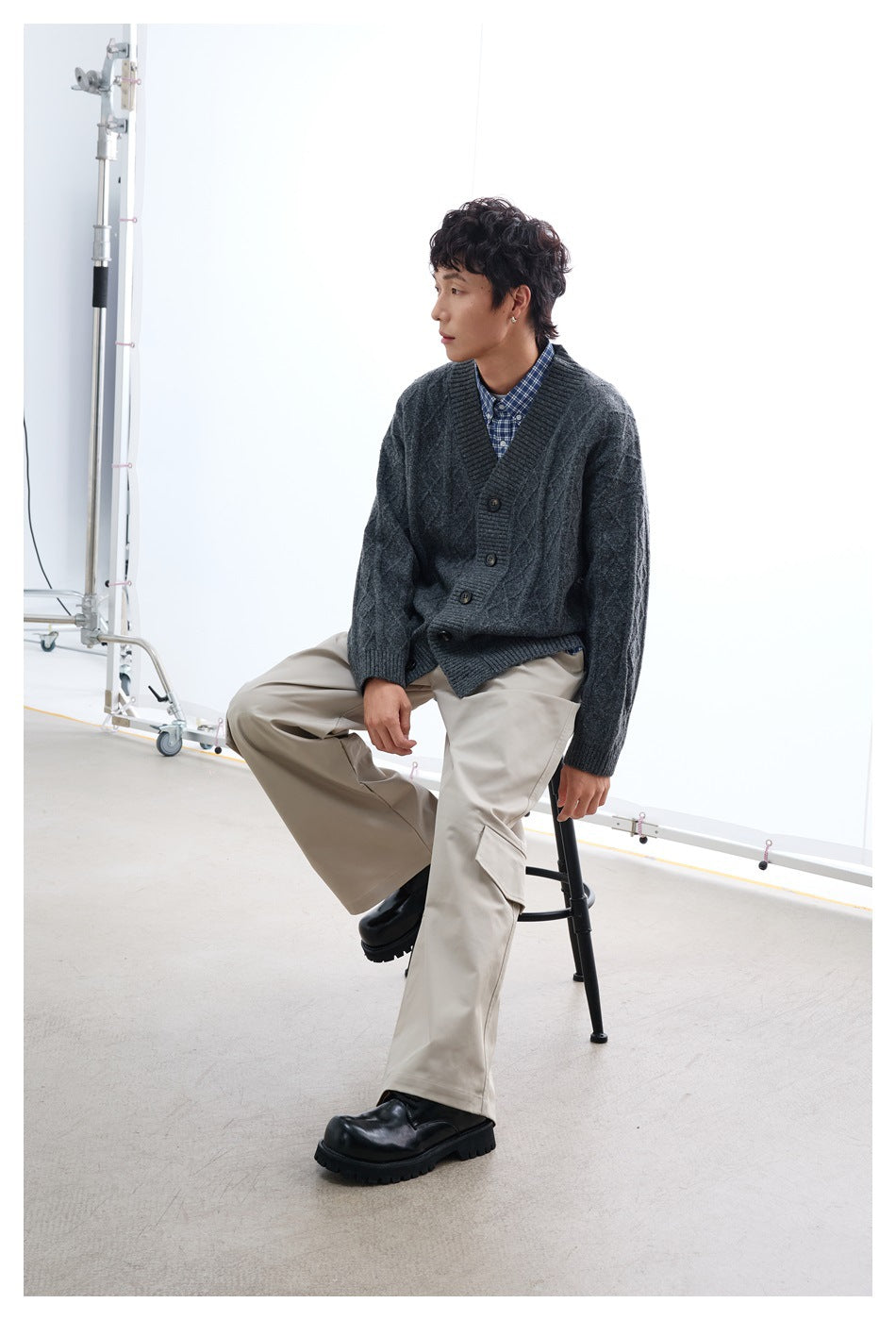 Seated model wearing a dark charcoal cable-knit button-front cardigan over a blue checked shirt.