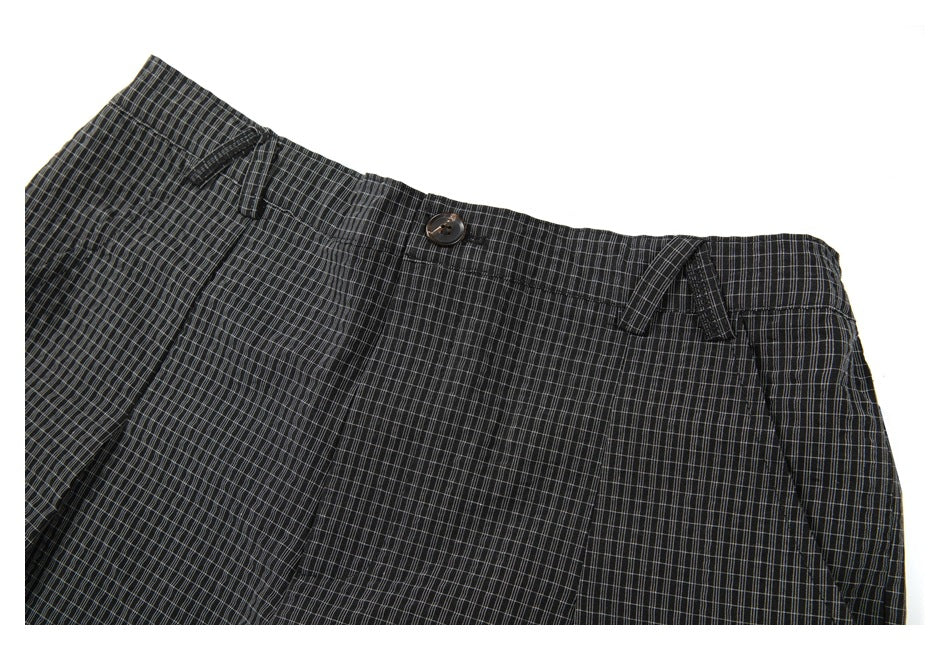 Angled close-up of the waistband, button, belt loops and pleats on dark gray plaid shorts.