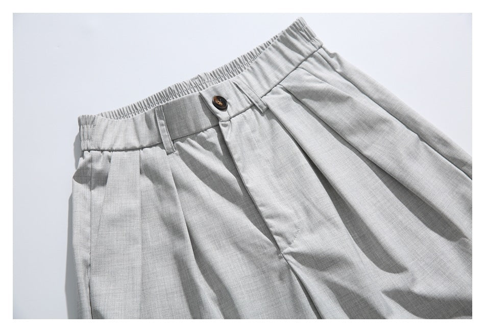 Close-up of light gray pleated trousers with an elasticized waistband, belt loop, and button.
