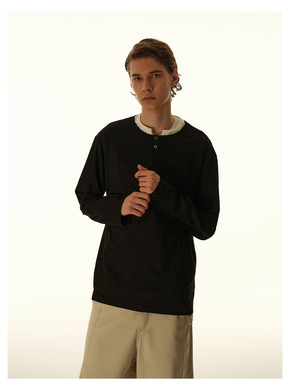Model wearing a black long-sleeve henley over a white shirt, with a short button placket and beige trousers.