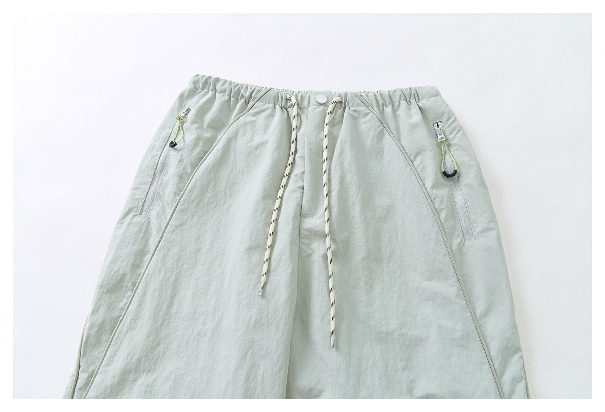 Front view of pale sage-gray pants with an elastic waist, patterned drawcord, curved piping, and zip pockets.