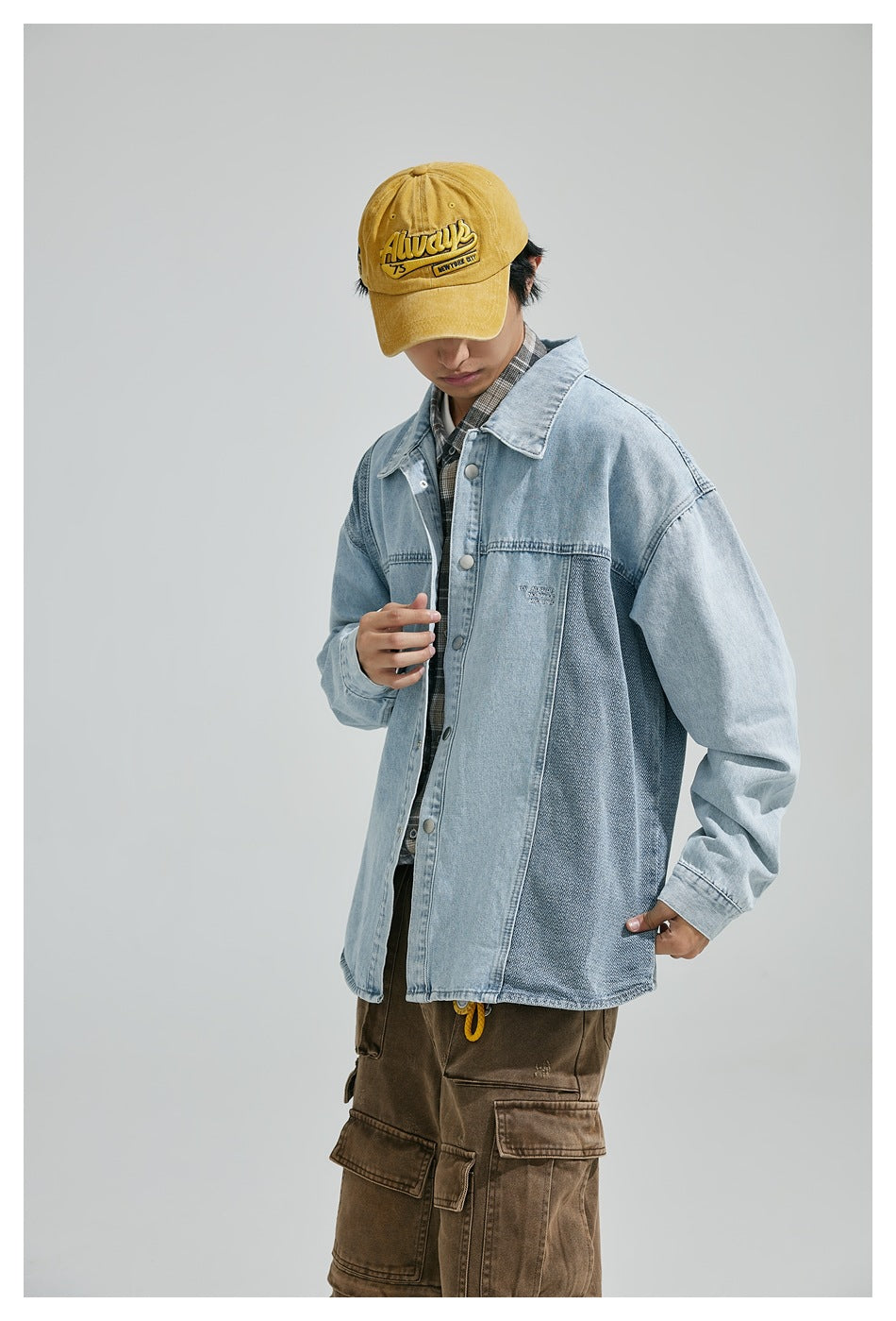 Model wearing a light-blue denim jacket with darker contrast panels, viewed from the front with a yellow cap and brown cargo