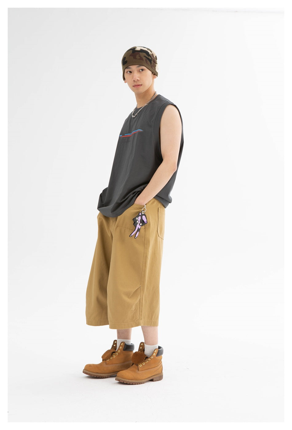 Model wearing a charcoal sleeveless top with a small colored graphic, tan wide-leg shorts, camo beanie and tan boots.
