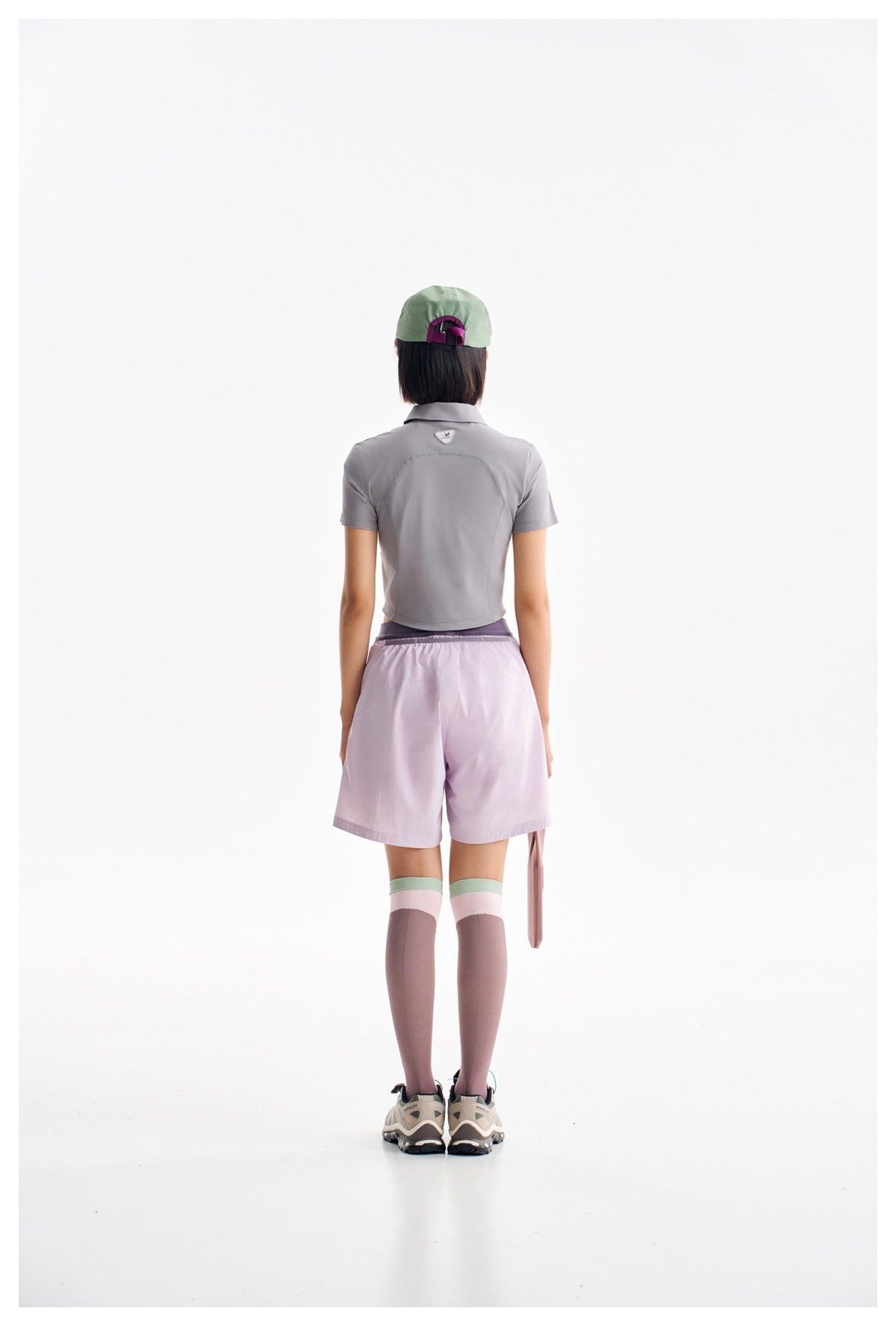 Rear view of a model wearing a gray cropped polo, lilac shorts, pink knee-high socks and a mint cap.