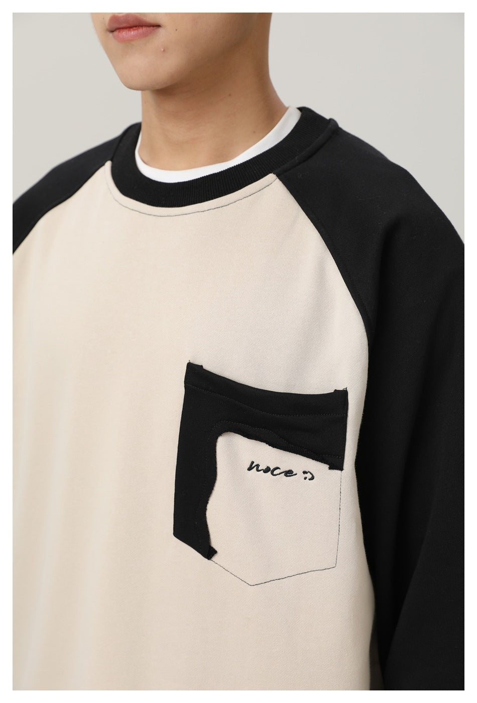 Close-up of a person wearing a cream sweatshirt with black raglan sleeves, a ribbed collar and a contrast chest pocket.