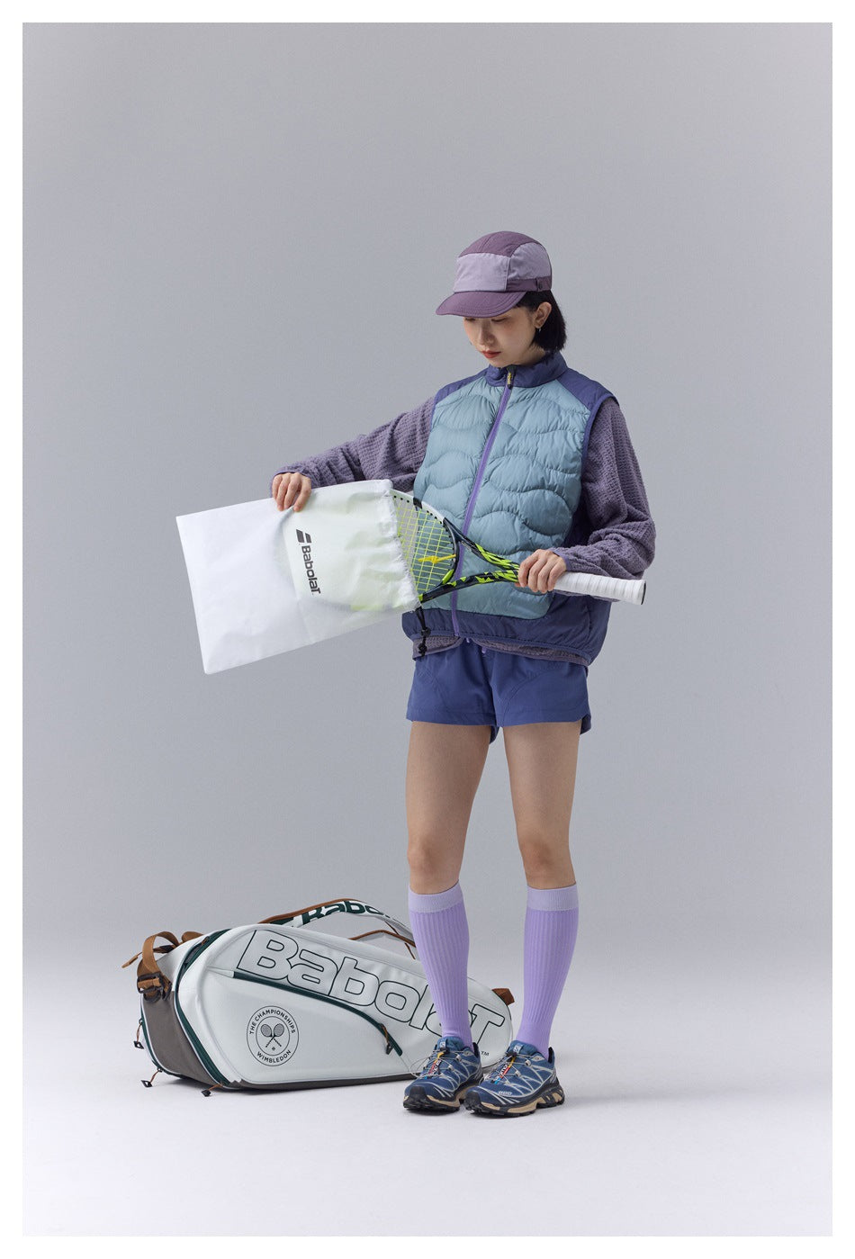 Model wearing a pale blue quilted vest with purple trim, purple textured top and blue shorts while holding a tennis racket.