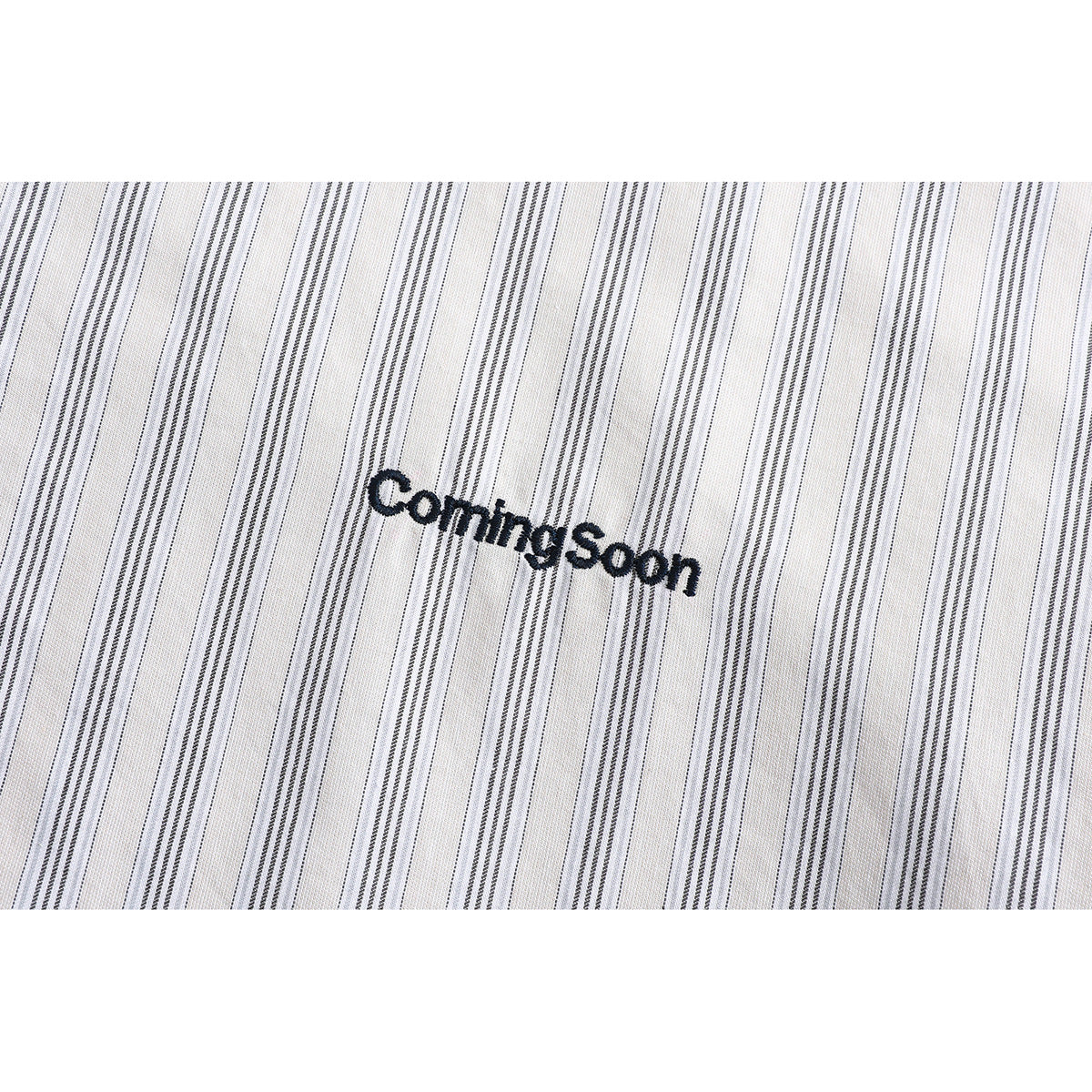 Close-up of thin gray stripes and black embroidered “ComingSoon” lettering on white shirt fabric.
