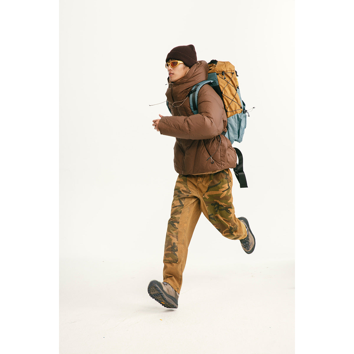 Model running in a brown hooded puffer jacket with camouflage pants and a hiking backpack.