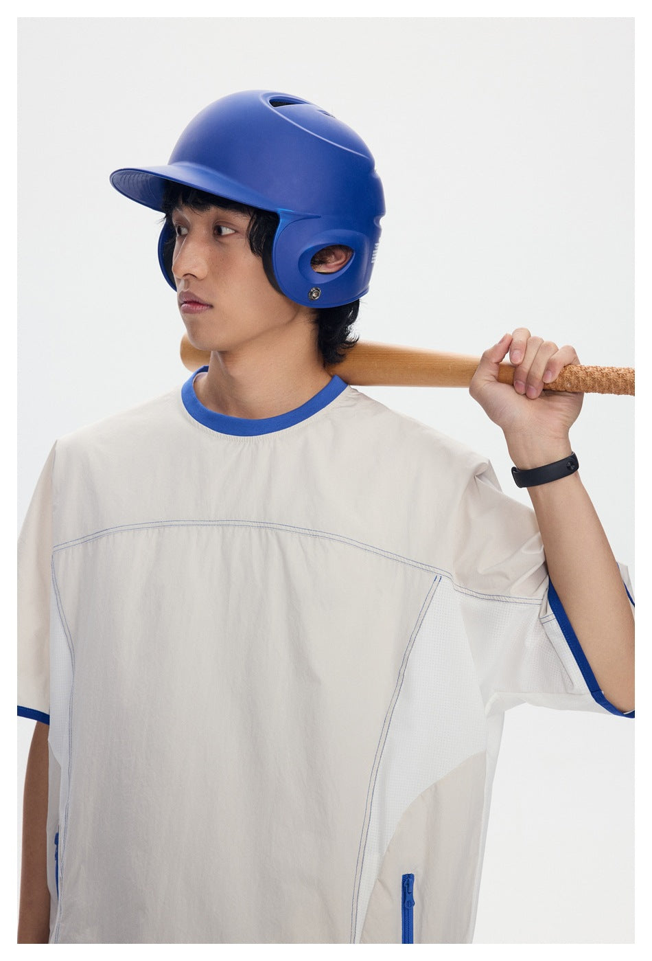 Model wearing an oversized cream shirt with blue trim and side zippers, holding a bat and wearing a blue helmet.