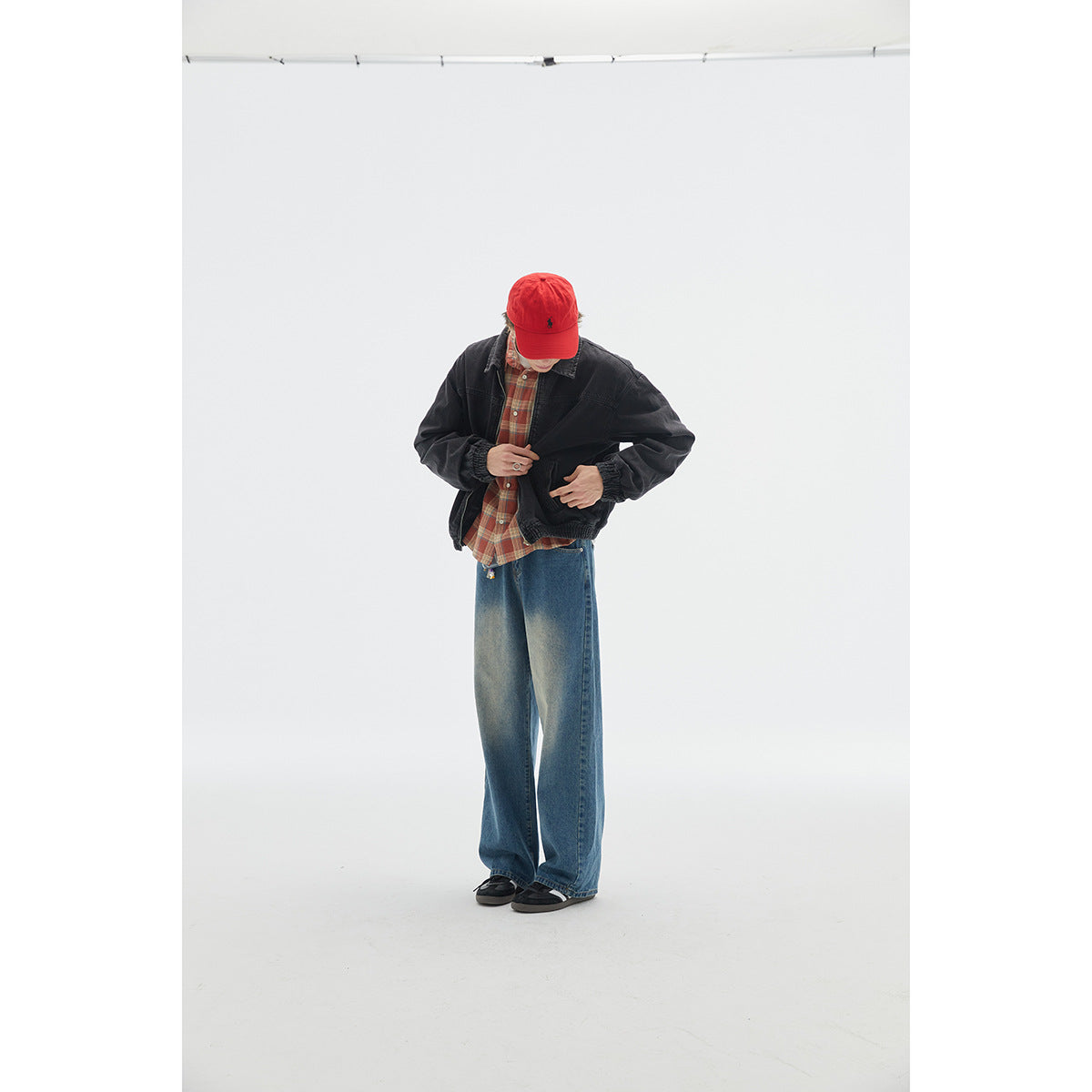 Model wearing a black jacket over a red plaid shirt and loose blue jeans, with a red cap.