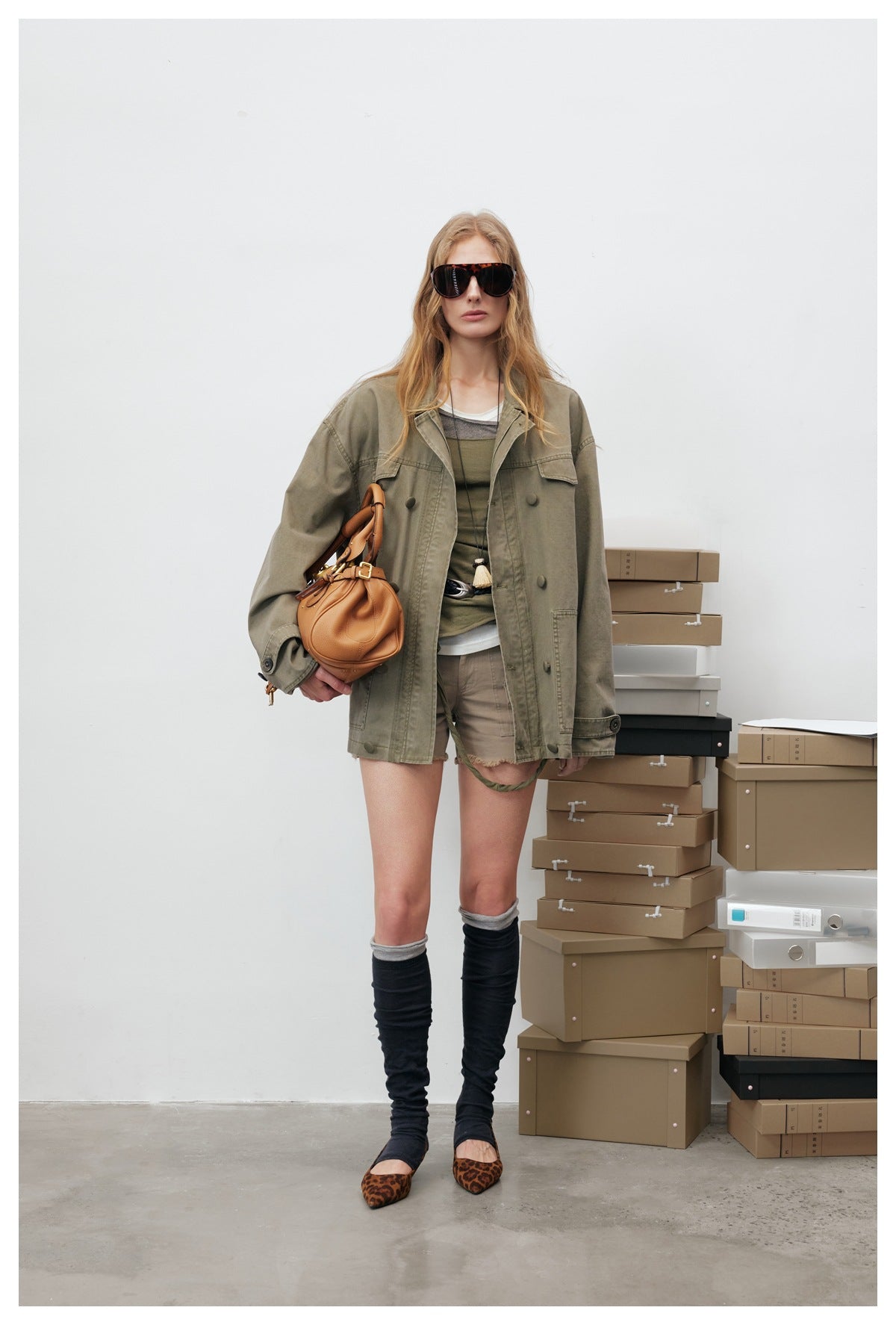 Woman wearing an open olive green oversized jacket with shorts, knee-high socks and a tan handbag, front view