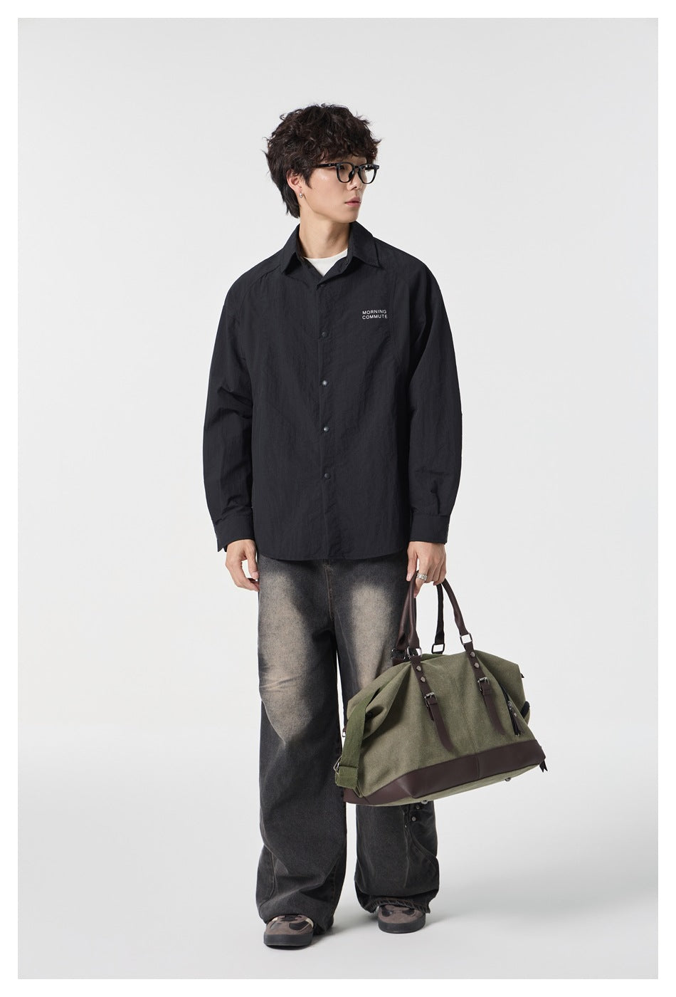 Full-length model wearing a black button-front overshirt and washed wide-leg jeans, holding an olive bag.