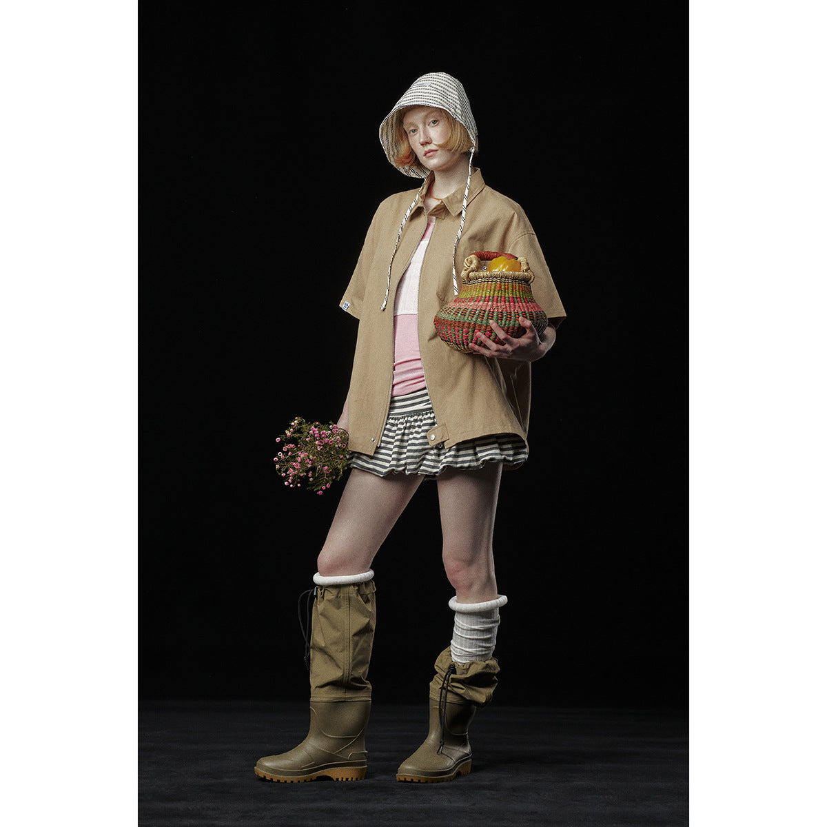 Model wearing a tan short-sleeve zip shirt over a pink top, striped skirt, patterned hood, and tall olive boots.