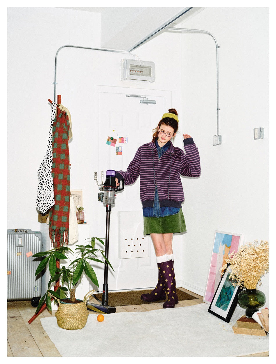 Model wearing a purple-and-black striped zip jacket over denim and a green skirt, shown in a bright room.