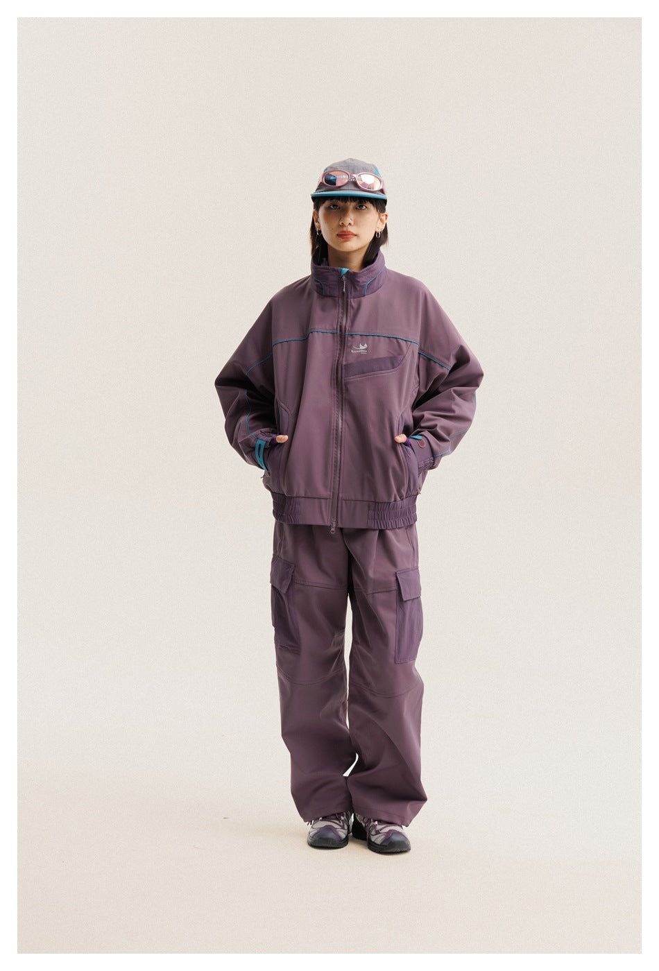 Model wearing a plum zip jacket with turquoise piping and matching cargo pants, standing with hands in the jacket pockets.