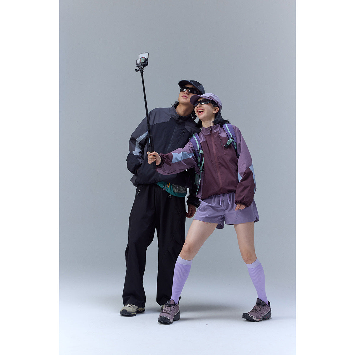 Two models pose with an action camera; one wears a dark jacket and trousers, the other a plum jacket and lilac shorts.