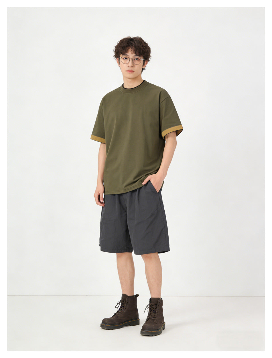 Model wearing an olive crew-neck T-shirt with mustard sleeve cuffs and charcoal shorts, shown full length.