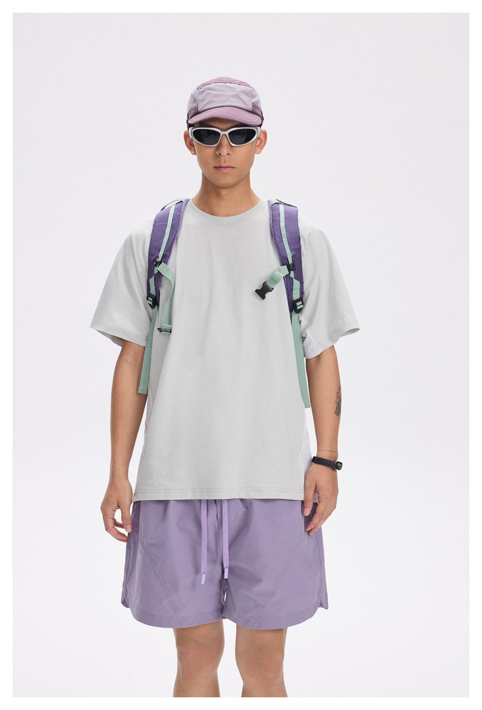 Front view of a model in a light gray short-sleeve T-shirt with purple shorts, cap, sunglasses and backpack straps